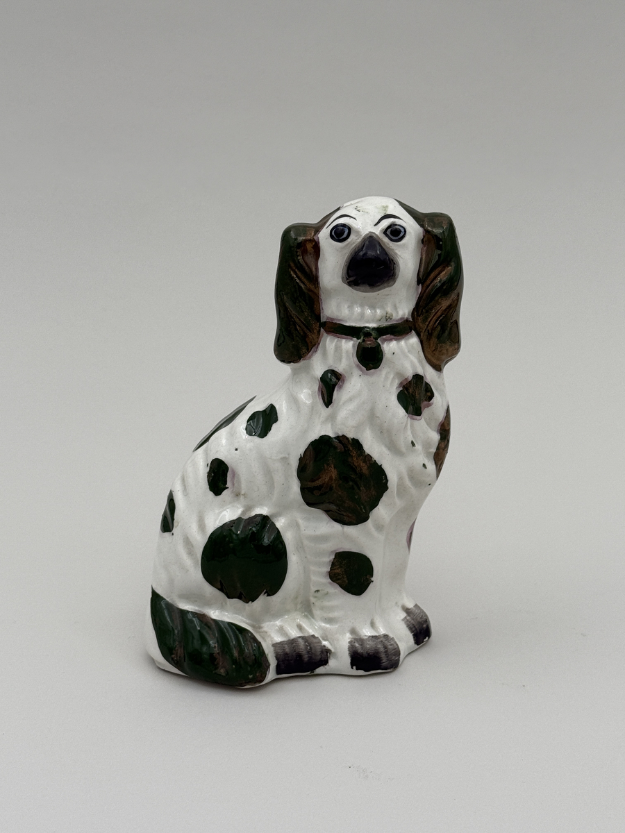 Figurin av porslin i form av sittande hund med halsband, glaserad, med överglasymålad dekor och förgyllning (förgyllningen mestadels bortnött).  
 