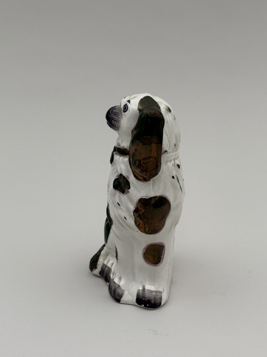 Figurin av porslin i form av sittande hund med halsband, glaserad, med överglasymålad dekor och förgyllning (förgyllningen mestadels bortnött).  
 