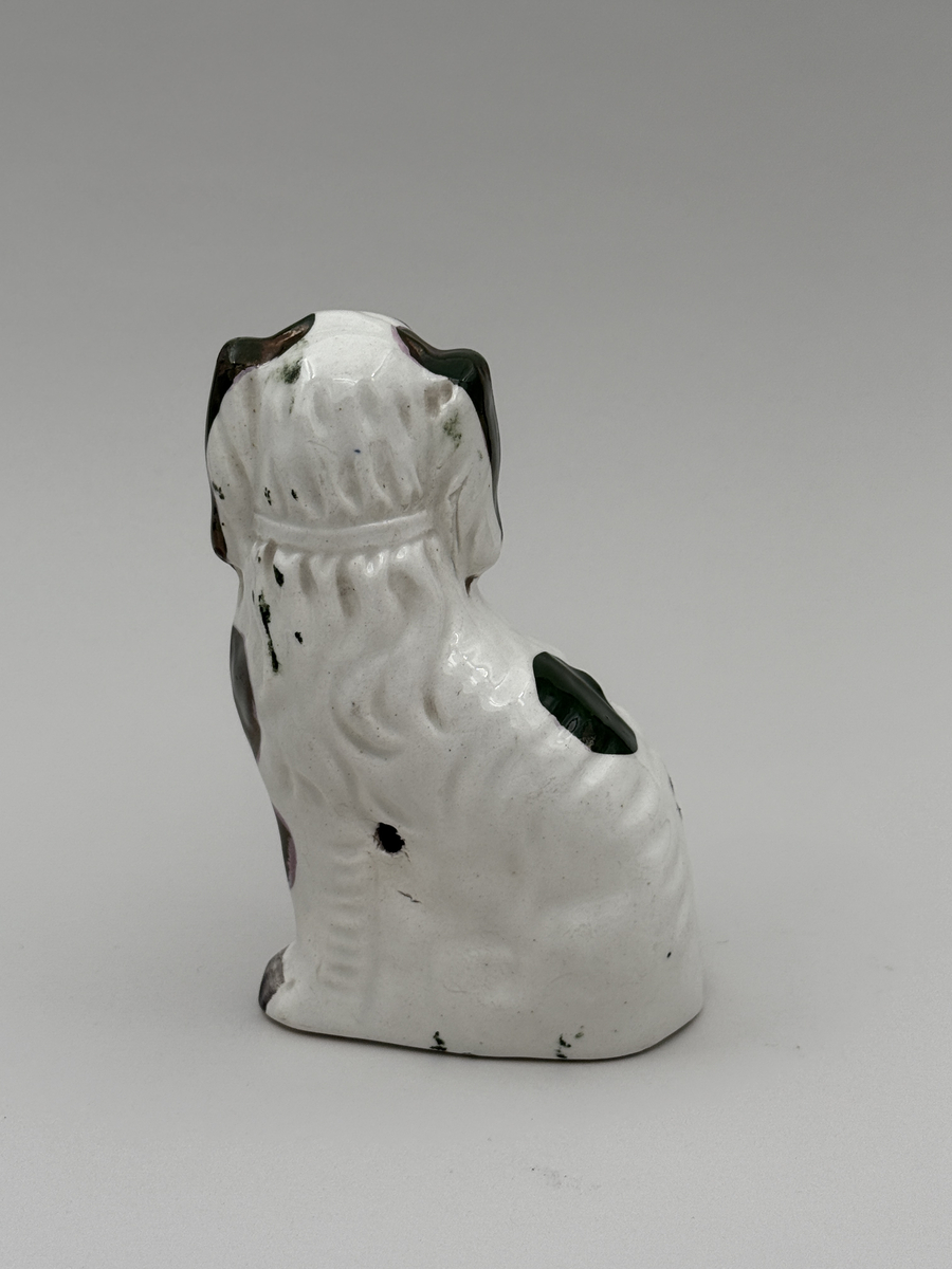 Figurin av porslin i form av sittande hund med halsband, glaserad, med överglasymålad dekor och förgyllning (förgyllningen mestadels bortnött).  
 