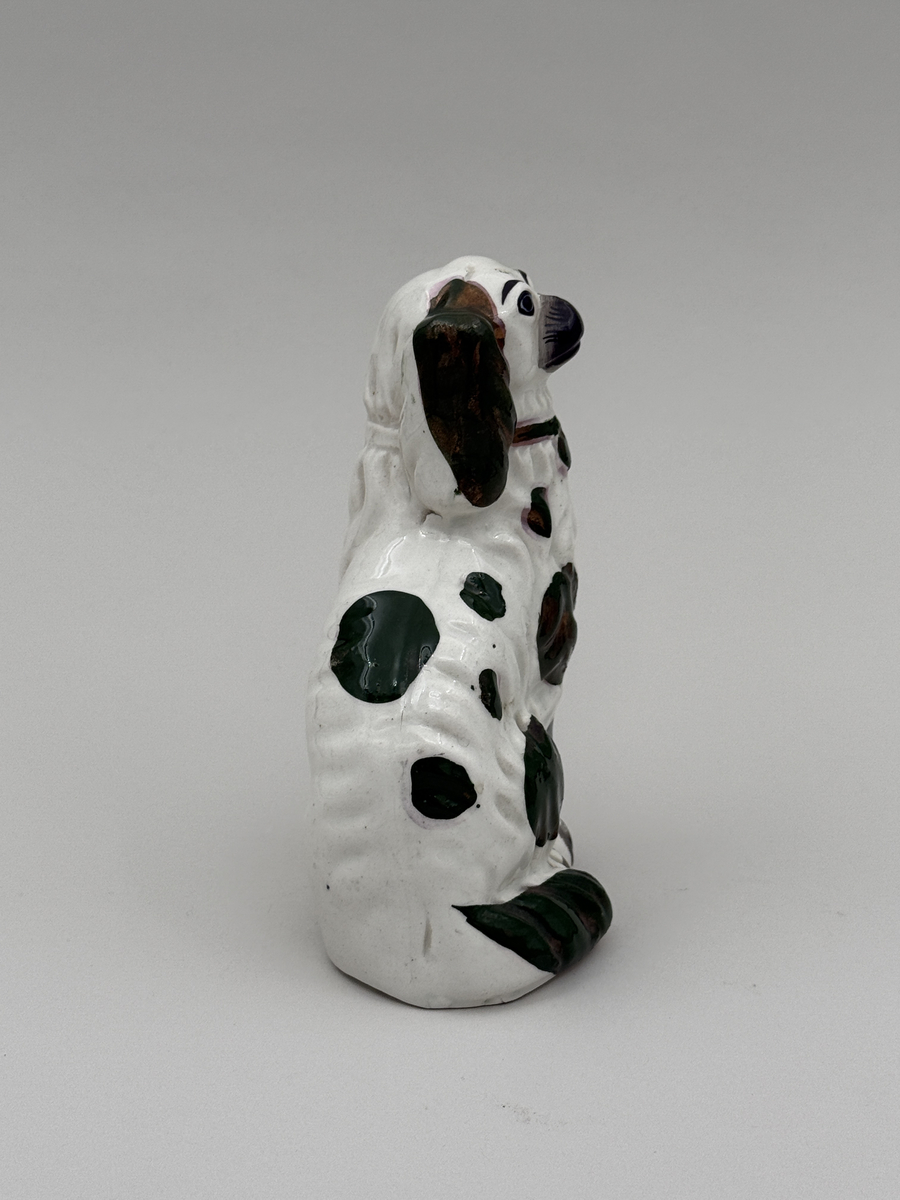 Figurin av porslin i form av sittande hund med halsband, glaserad, med överglasymålad dekor och förgyllning (förgyllningen mestadels bortnött).  
 