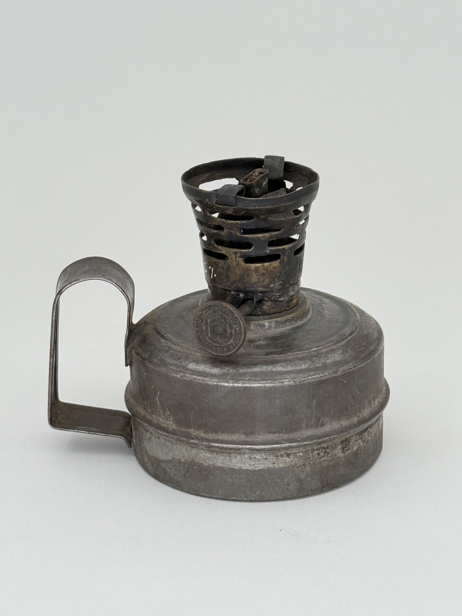 Oljelampa med rund behållare av bleckplåt, handtag med hål för upphängning. Brännare av mässing med liten ratt för att mata upp veken. Ratten stämplad "Arvid Böhlmark Stockholm" samt med firmans logga: en antik oljelampa. 