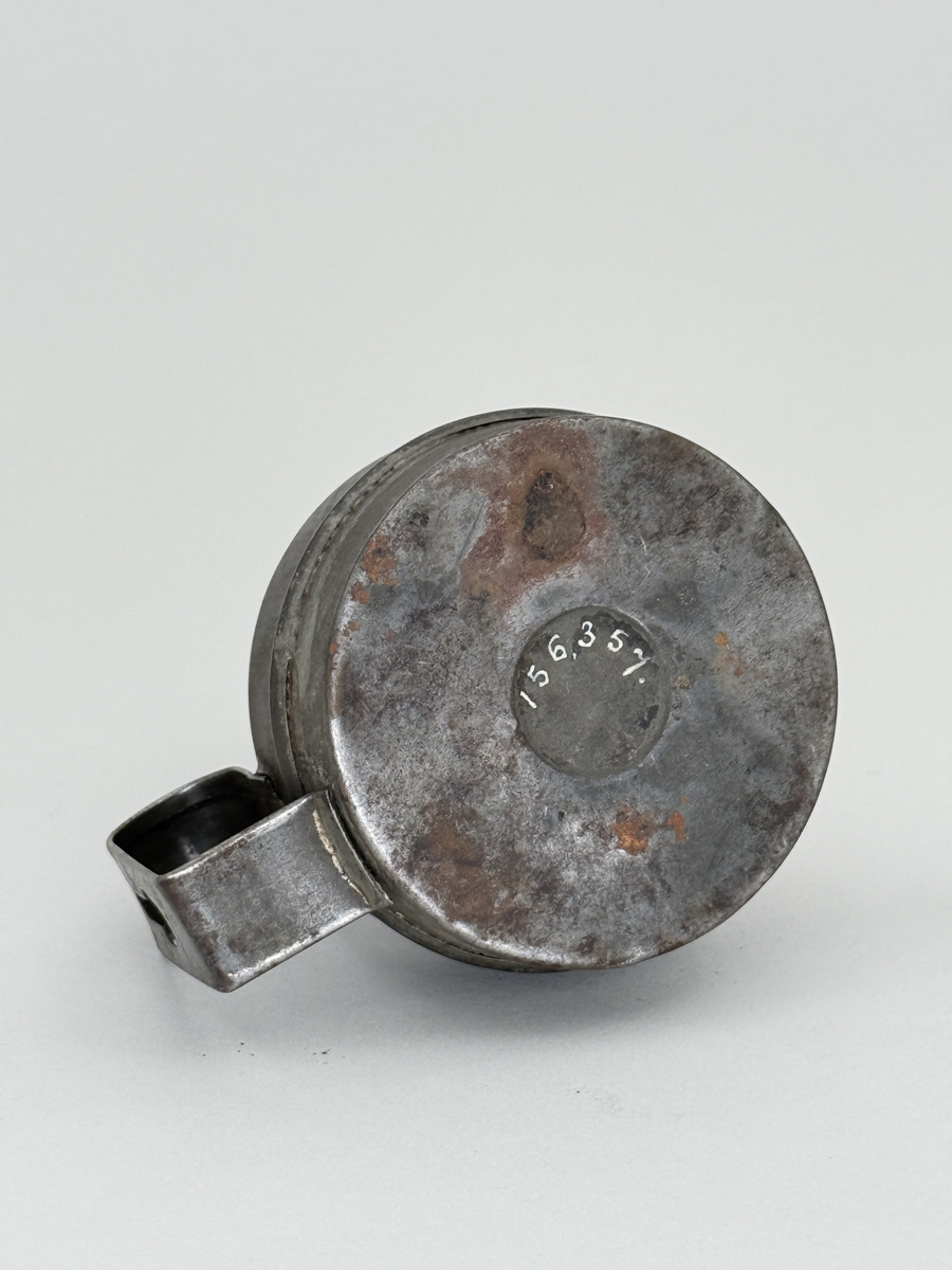 Oljelampa med rund behållare av bleckplåt, handtag med hål för upphängning. Brännare av mässing med liten ratt för att mata upp veken. Ratten stämplad "Arvid Böhlmark Stockholm" samt med firmans logga: en antik oljelampa. 