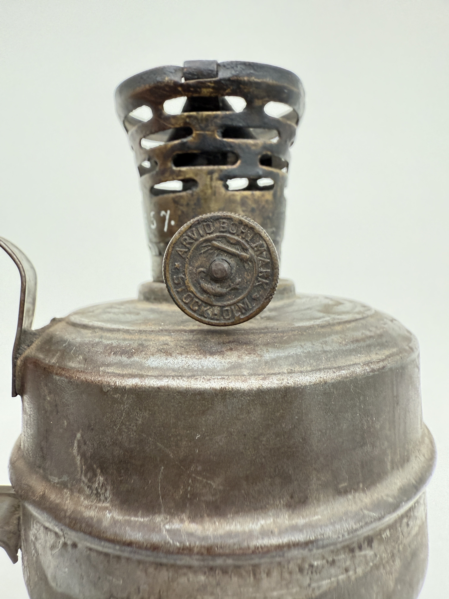 Oljelampa med rund behållare av bleckplåt, handtag med hål för upphängning. Brännare av mässing med liten ratt för att mata upp veken. Ratten stämplad "Arvid Böhlmark Stockholm" samt med firmans logga: en antik oljelampa. 
