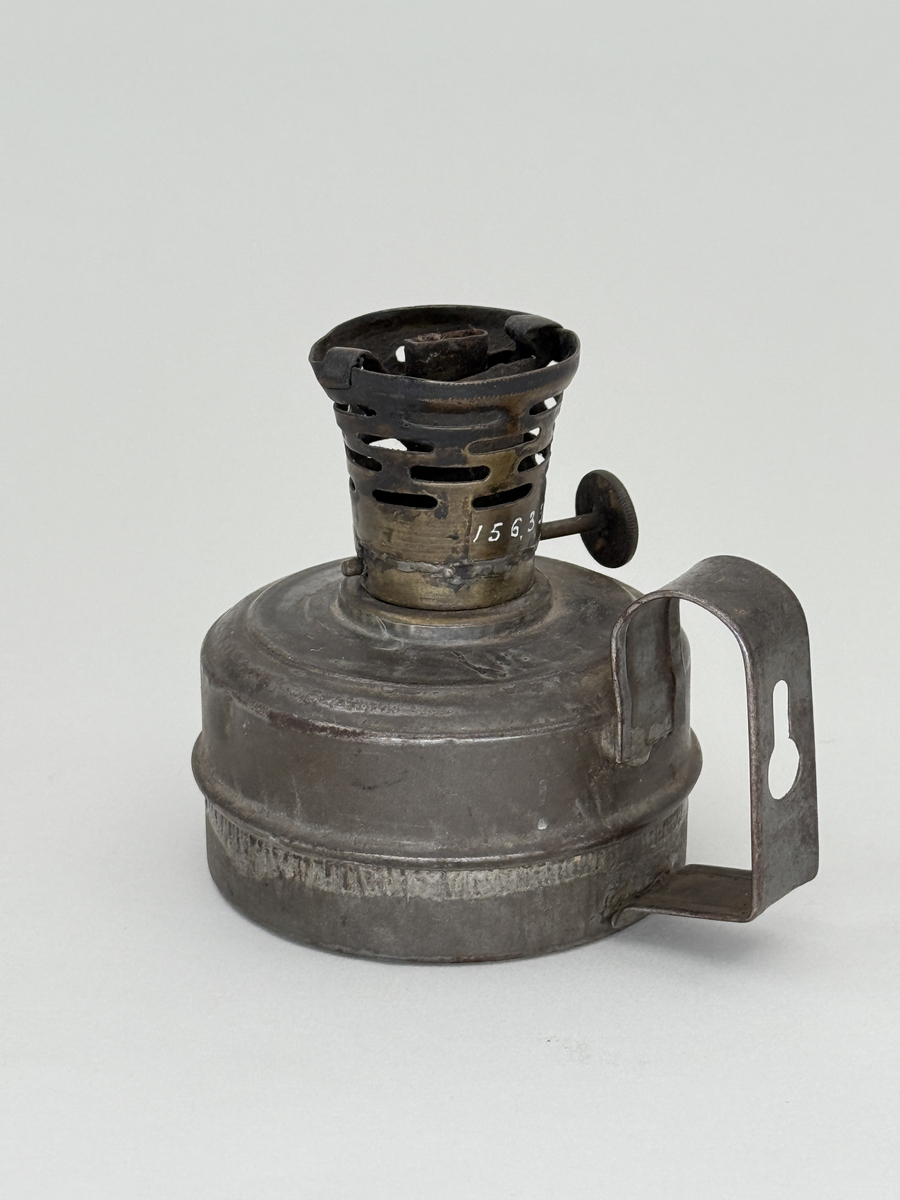 Oljelampa med rund behållare av bleckplåt, handtag med hål för upphängning. Brännare av mässing med liten ratt för att mata upp veken. Ratten stämplad "Arvid Böhlmark Stockholm" samt med firmans logga: en antik oljelampa. 