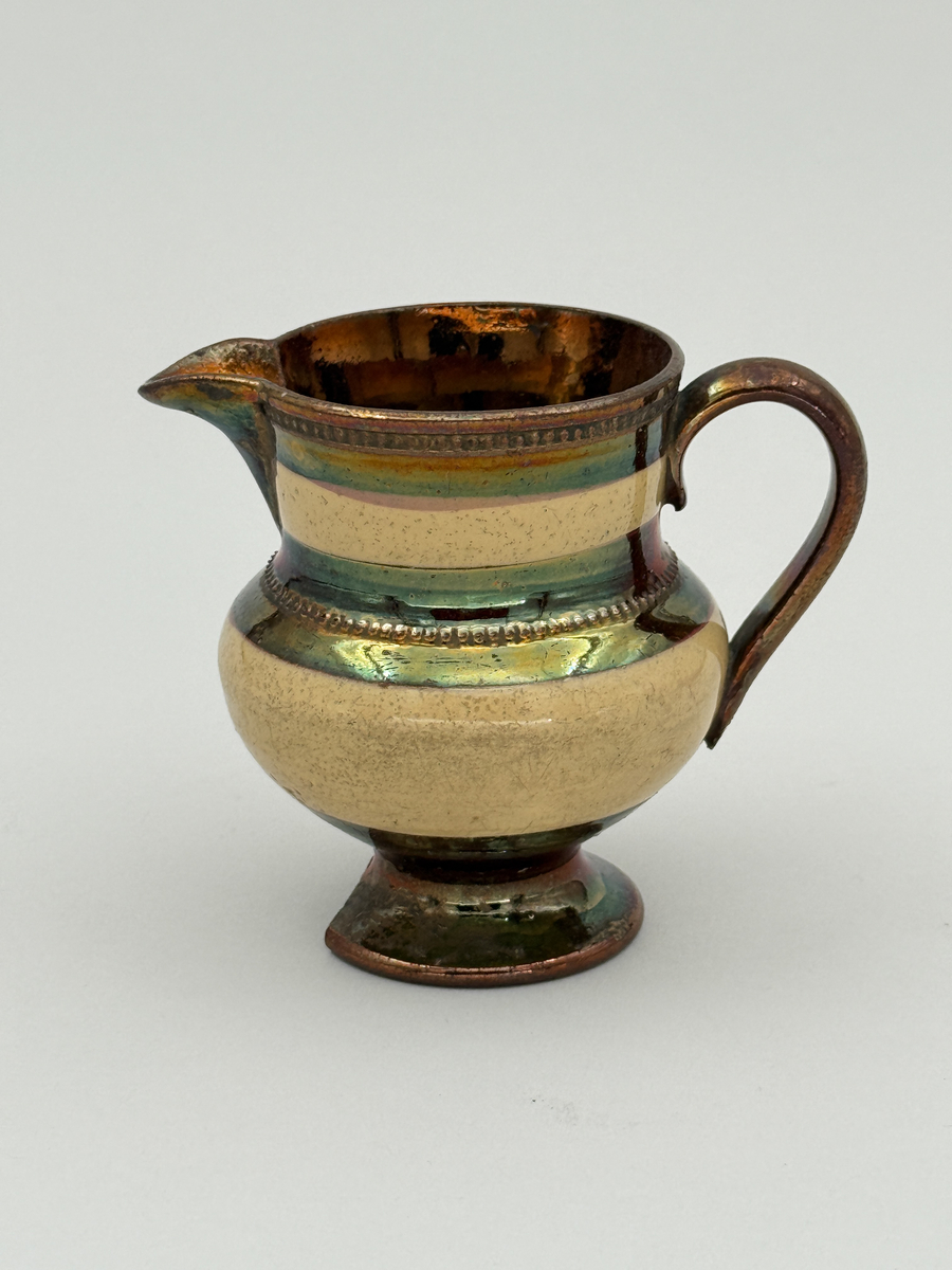 Gräddkanna av porslin, bukig form på indragen hög fot samt med utsvängd pip. Spräckligt gul glasyr, lysterglasyr samt bronsering. Dekorerad med pärlstav.