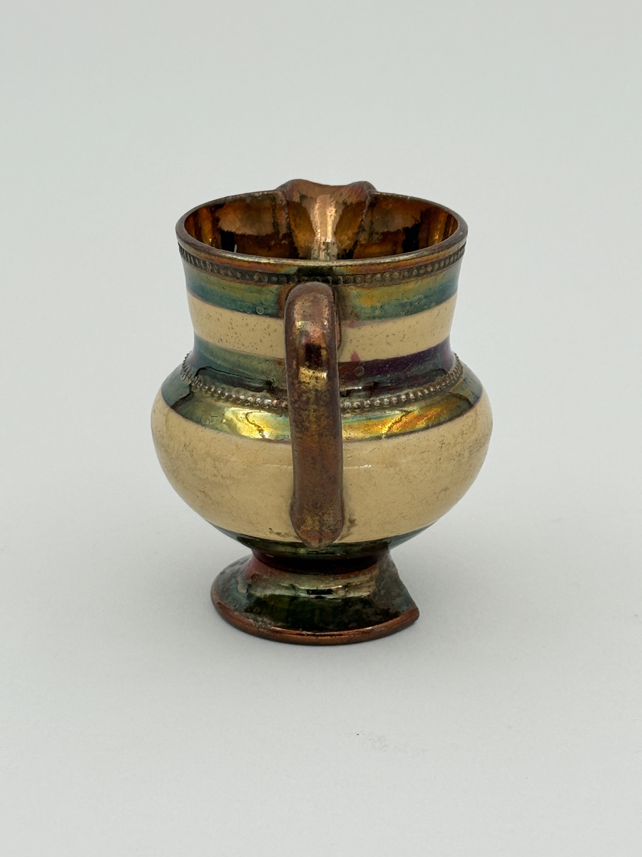 Gräddkanna av porslin, bukig form på indragen hög fot samt med utsvängd pip. Spräckligt gul glasyr, lysterglasyr samt bronsering. Dekorerad med pärlstav.