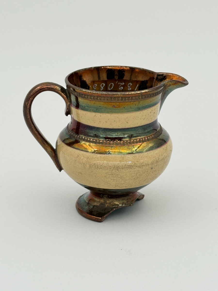 Gräddkanna av porslin, bukig form på indragen hög fot samt med utsvängd pip. Spräckligt gul glasyr, lysterglasyr samt bronsering. Dekorerad med pärlstav.