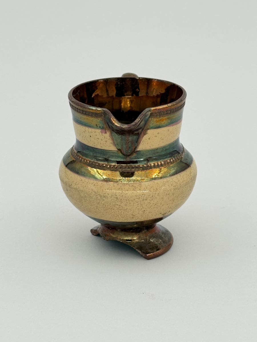 Gräddkanna av porslin, bukig form på indragen hög fot samt med utsvängd pip. Spräckligt gul glasyr, lysterglasyr samt bronsering. Dekorerad med pärlstav.