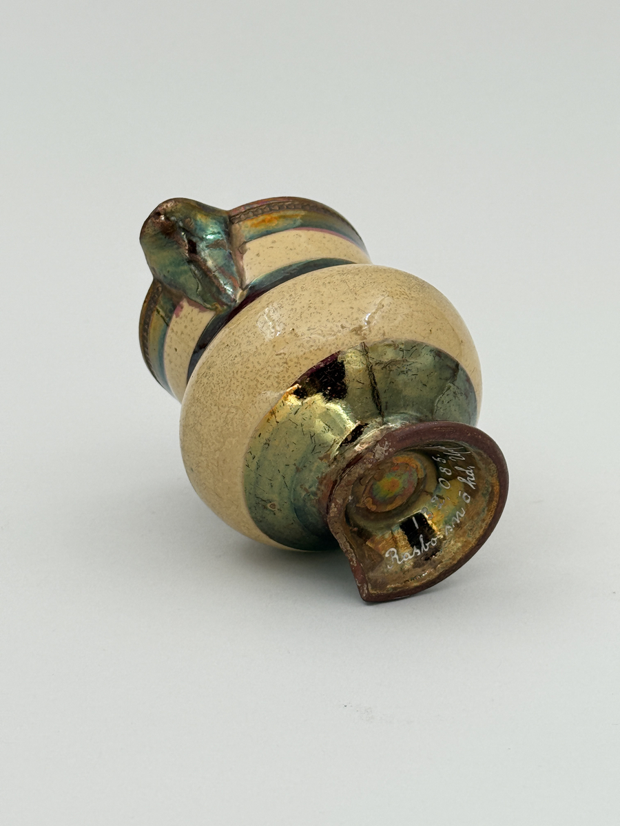 Gräddkanna av porslin, bukig form på indragen hög fot samt med utsvängd pip. Spräckligt gul glasyr, lysterglasyr samt bronsering. Dekorerad med pärlstav.