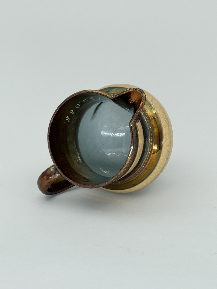 Gräddkanna av porslin, bukig form på indragen hög fot samt med utsvängd pip. Spräckligt gul glasyr, lysterglasyr samt bronsering. Dekorerad med pärlstav.