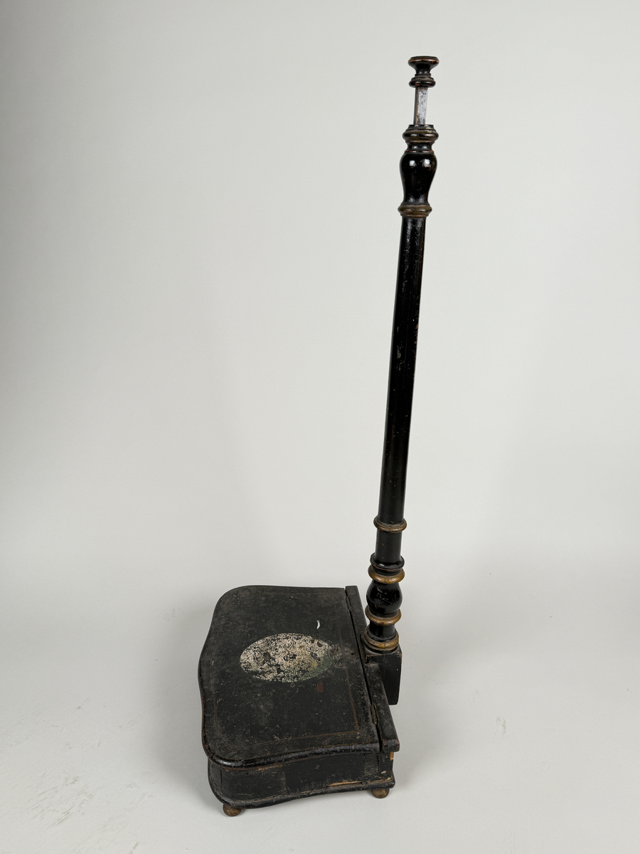 Spottlåda av svärtat trä, delvis fanerad, på kulfötter.  Lock med gångjärn, dekorerat med ljus medaljong. Profilsvarvat skaft fäst vid lådans bakre kant, mekanisk öppning genom att man trycker på knappen överst på skaftet. 