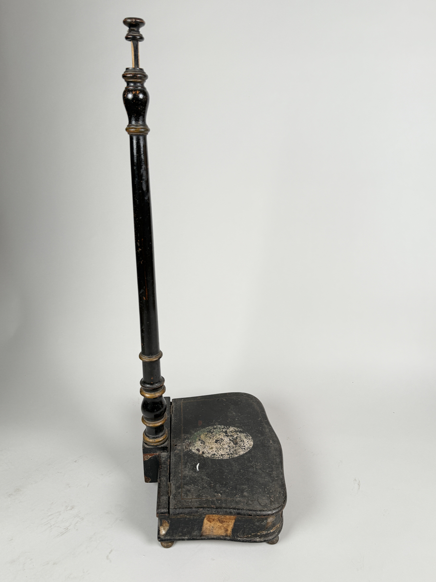 Spottlåda av svärtat trä, delvis fanerad, på kulfötter.  Lock med gångjärn, dekorerat med ljus medaljong. Profilsvarvat skaft fäst vid lådans bakre kant, mekanisk öppning genom att man trycker på knappen överst på skaftet. 