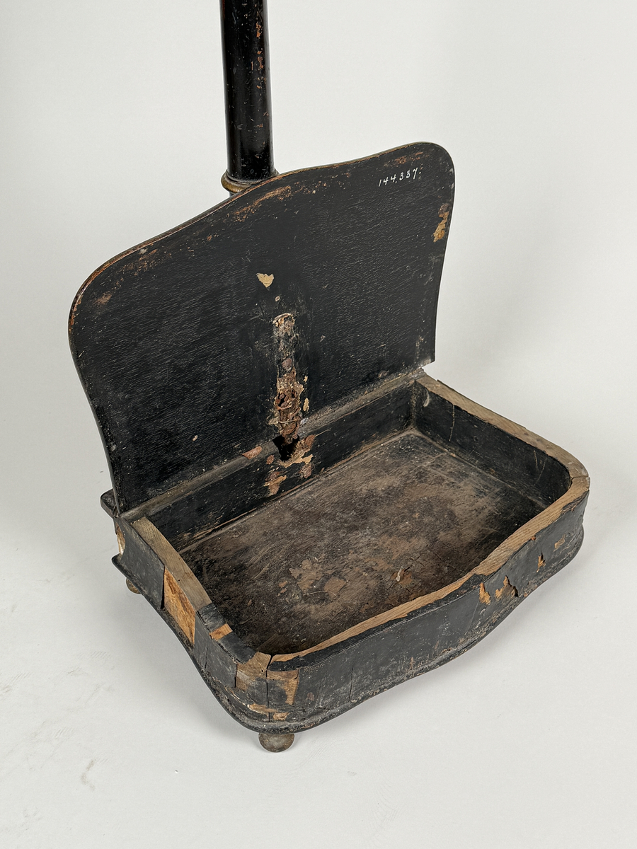 Spottlåda av svärtat trä, delvis fanerad, på kulfötter.  Lock med gångjärn, dekorerat med ljus medaljong. Profilsvarvat skaft fäst vid lådans bakre kant, mekanisk öppning genom att man trycker på knappen överst på skaftet. 