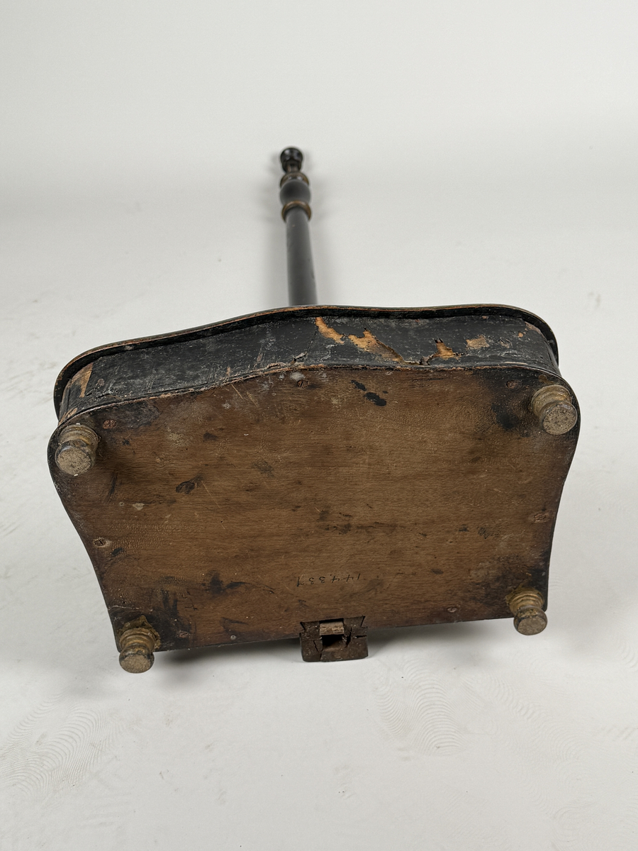 Spottlåda av svärtat trä, delvis fanerad, på kulfötter.  Lock med gångjärn, dekorerat med ljus medaljong. Profilsvarvat skaft fäst vid lådans bakre kant, mekanisk öppning genom att man trycker på knappen överst på skaftet. 