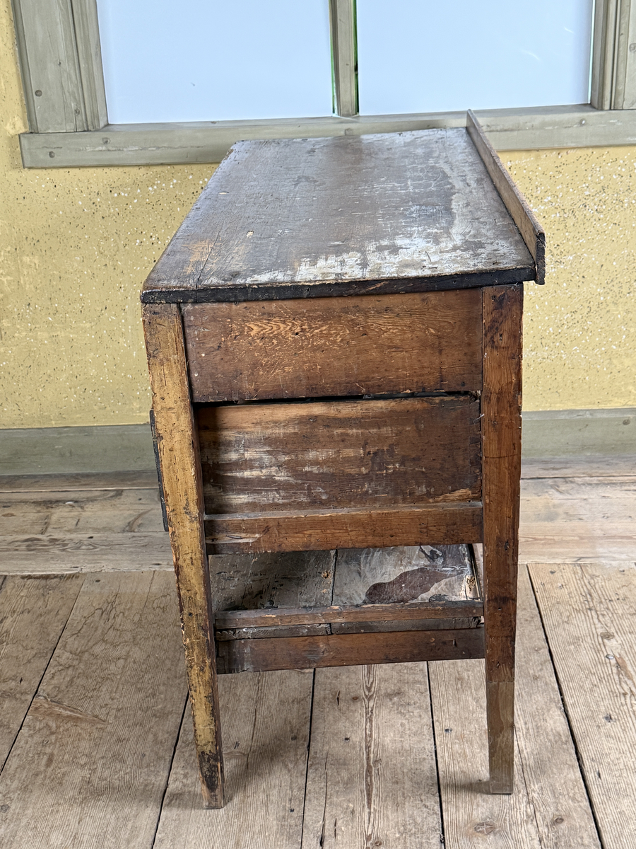 Avlastningsbord med rektangulär form, bemålad i beige-gult. Trågkant i skivans bakre kant. Övre lådvarvet med tre lådor, det undre lådvarvet med en låda. Längst ner en hylla. Dragknoppar av mässing, några saknas.