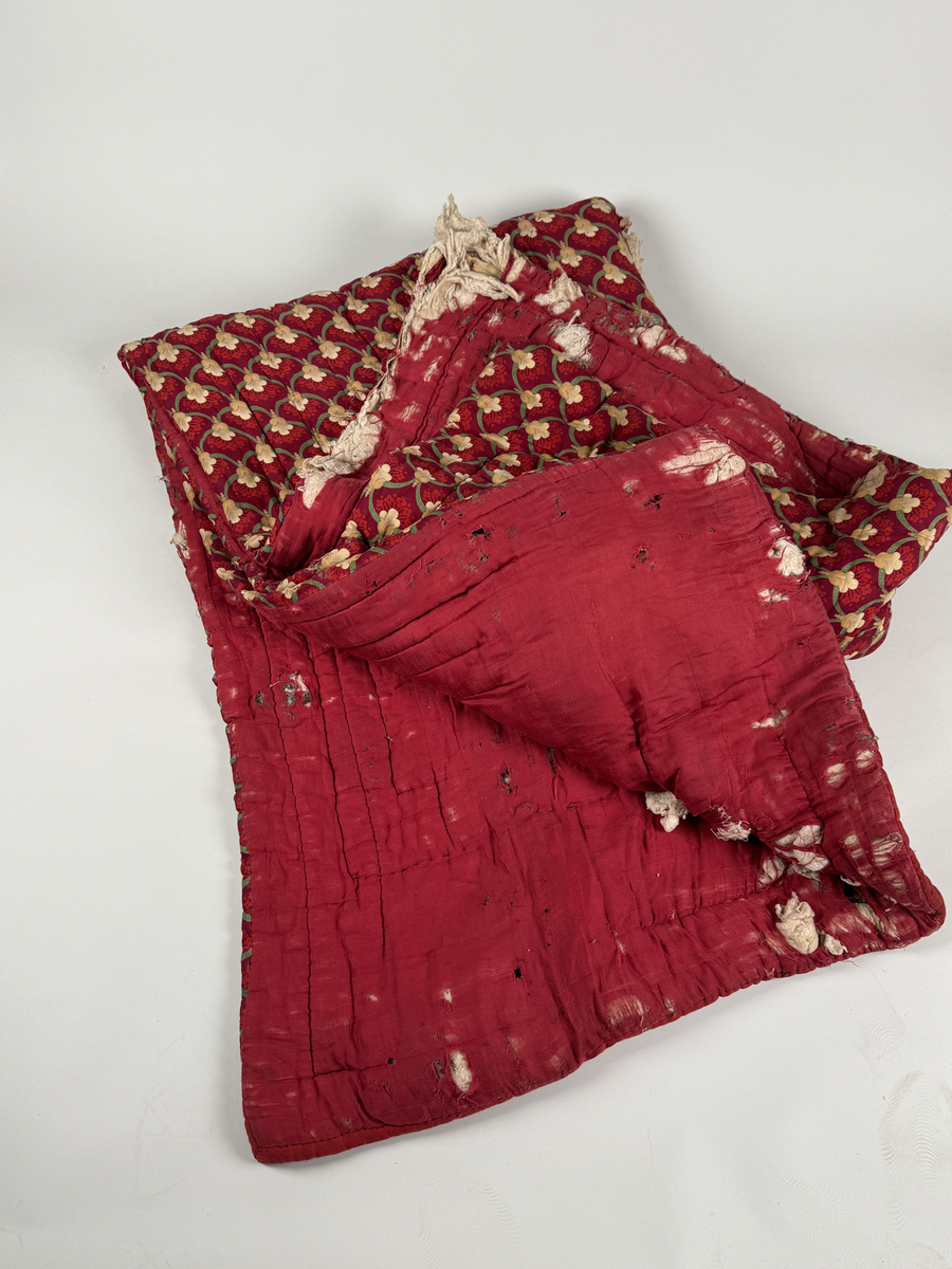 Vadderat täcke av bomull. Ovansidan med geometriskt mönster av blommor i grönt och gult mot rött, undersidan röd. 
