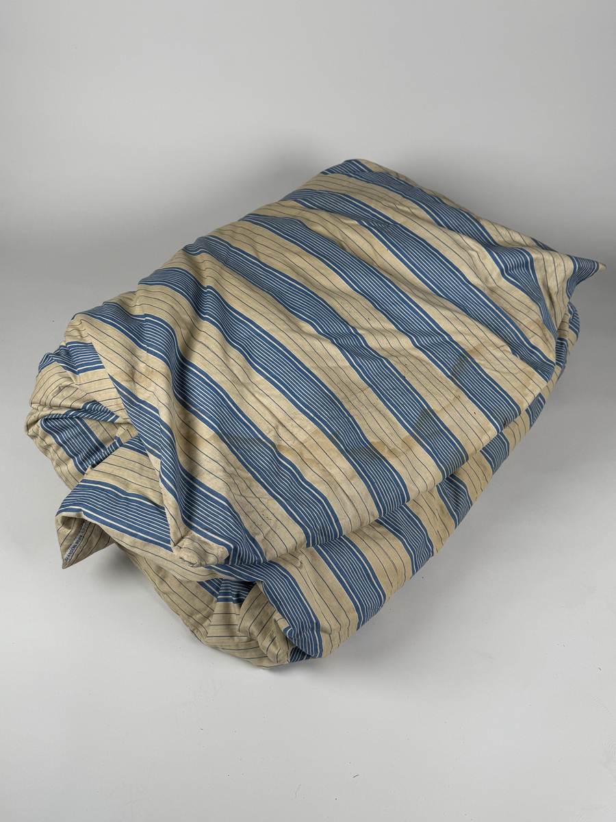 Bolster av bomull vävt i spetskypert, randigt i oblekt och mellanblått. Stoppat med dun. 
Gåva genom Leif Johansson.
