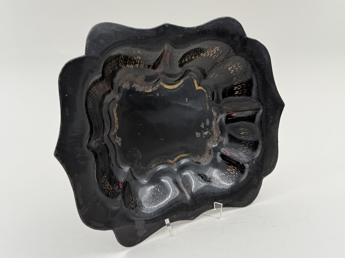 Svartlackerad brödkorg av plåt. Konturerad form med spår av förgylld dekor invändigt.