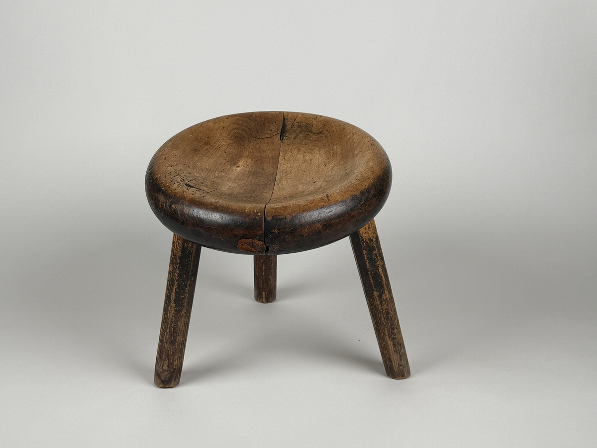 Pall med skålad sits av björk, åttkantiga ben av furu. Benen med ojämn längd så pallen lutar. Sitsen sprucken samt lagad med läderbitar och nubb. 