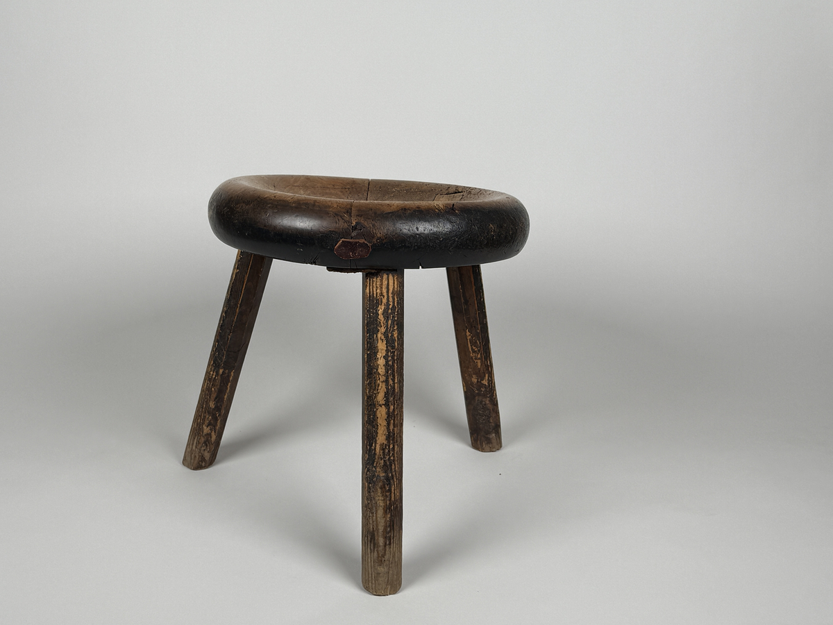 Pall med skålad sits av björk, åttkantiga ben av furu. Benen med ojämn längd så pallen lutar. Sitsen sprucken samt lagad med läderbitar och nubb. 