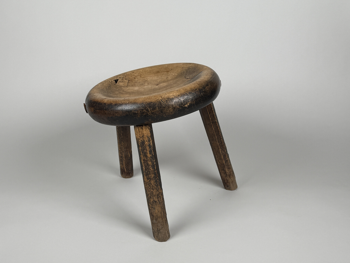 Pall med skålad sits av björk, åttkantiga ben av furu. Benen med ojämn längd så pallen lutar. Sitsen sprucken samt lagad med läderbitar och nubb. 