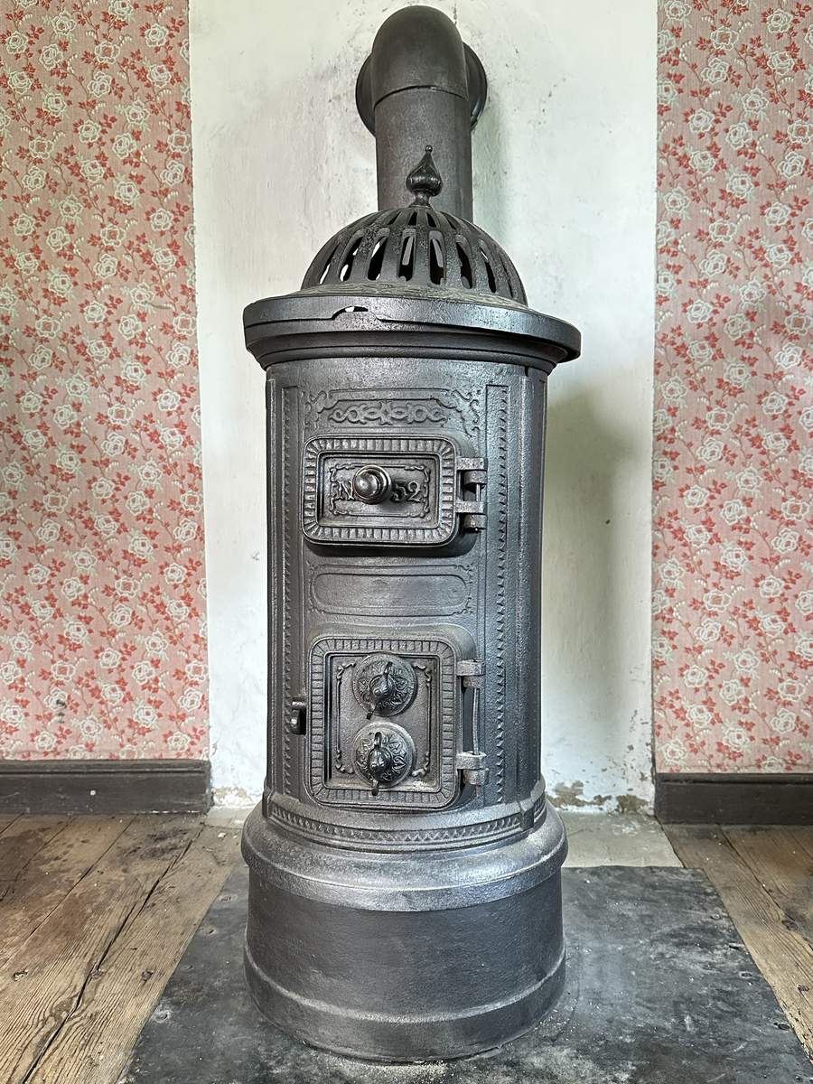 Cylindrisk gjutjärnskamin med spisringar och löstagbar, genombruten krona. Dekor av stiliserade bladrankor i godset. Vedlucka med spiralhandtag, asklucka med två dragventiler reglerade med vingmuttrar. Kaminen kopplad till rund trumma i bakkant. Kronan skadad i kanten. 