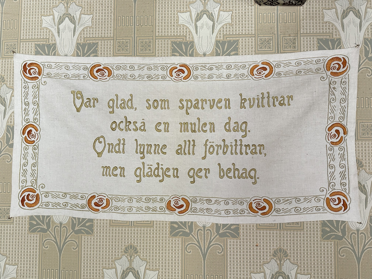 Väggbonad i vit linnelärft. Broderi i pärlgarn, stjälksöm och plattsöm (klumpsöm). Bård runt om med vågmönster i ljusgrönt, blommor i orange och rött. 