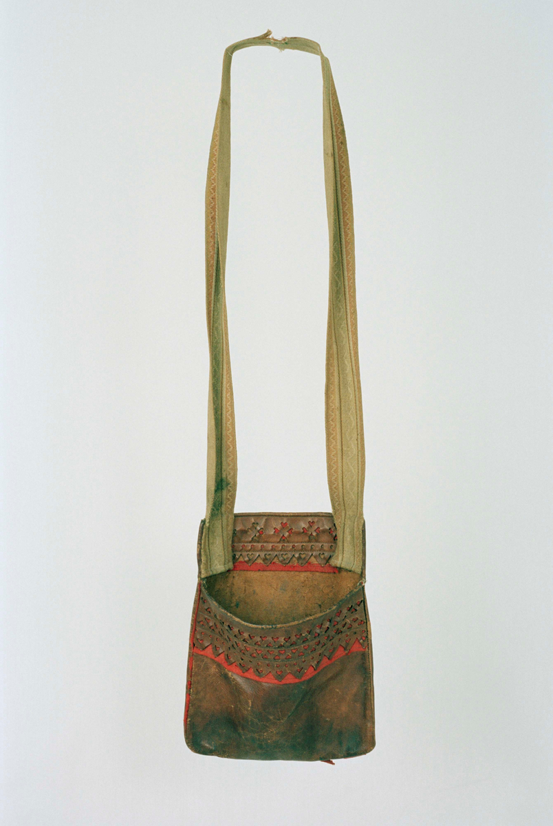Väska av brunt skinn, enkel form likt en ficka med axelrem. På framsidan genombrutet hjärtformat mönster. I sömmen runt om samt under dekoren rött ylletyg. Axelrem av textil i brunt, gult och grönt mot vit botten, kraftigt blekt. Axelremen förstärkt med sekundärt material vid slitskada mitt på bandet. Förstärkning gjord i samband med konservering. Vissa röda yllepartiker stabiliserade med infärgad organsa vid konservering.