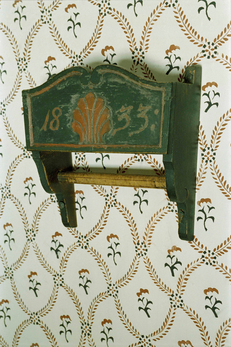 Handklähylla av furu, målad mörkgrön. Formad som en öppen låda med sidor och botten. Förlängda sidogavlar, nedtill profilerade, med instucken hängstång. Framsidan med snidad rocaillesnäcka i rött samt årtalet "1833".