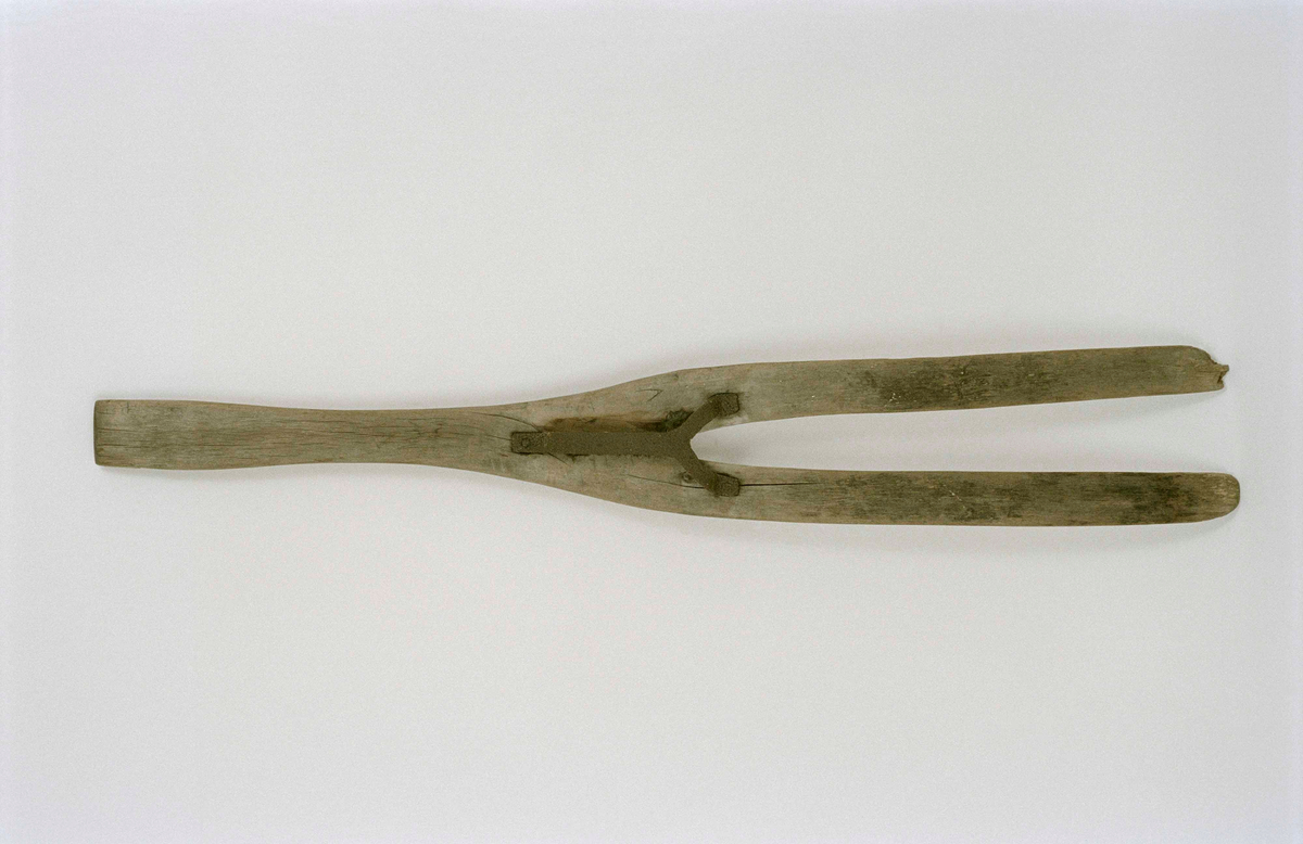 Dynggrep av björkträ. Platt, gaffelformig, två platta klor. På översidans mitt ett fastnitat Y-format handtag av järn.