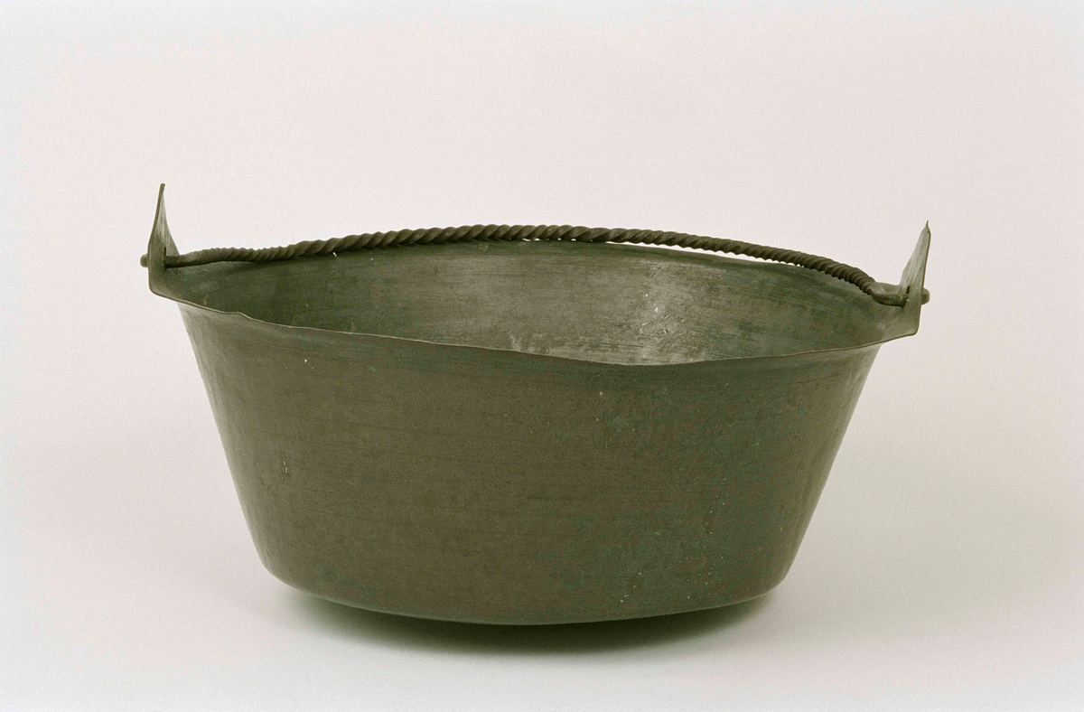 Kittel av kopparplåt, cylindrisk med utvikt mynningskant, öron i form av tringelformade flikar. Spiralvriden tvärgrepe. Spår av förtenning på insidan. Ristade linjer utmed kittelns utsida.