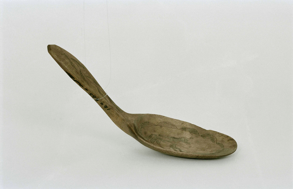 Sked av björk med ristad ornering i form av hästliknande djur,  ifylld med svart färg genom en teknik kalla kolrosning. Skaftänden avfasad.