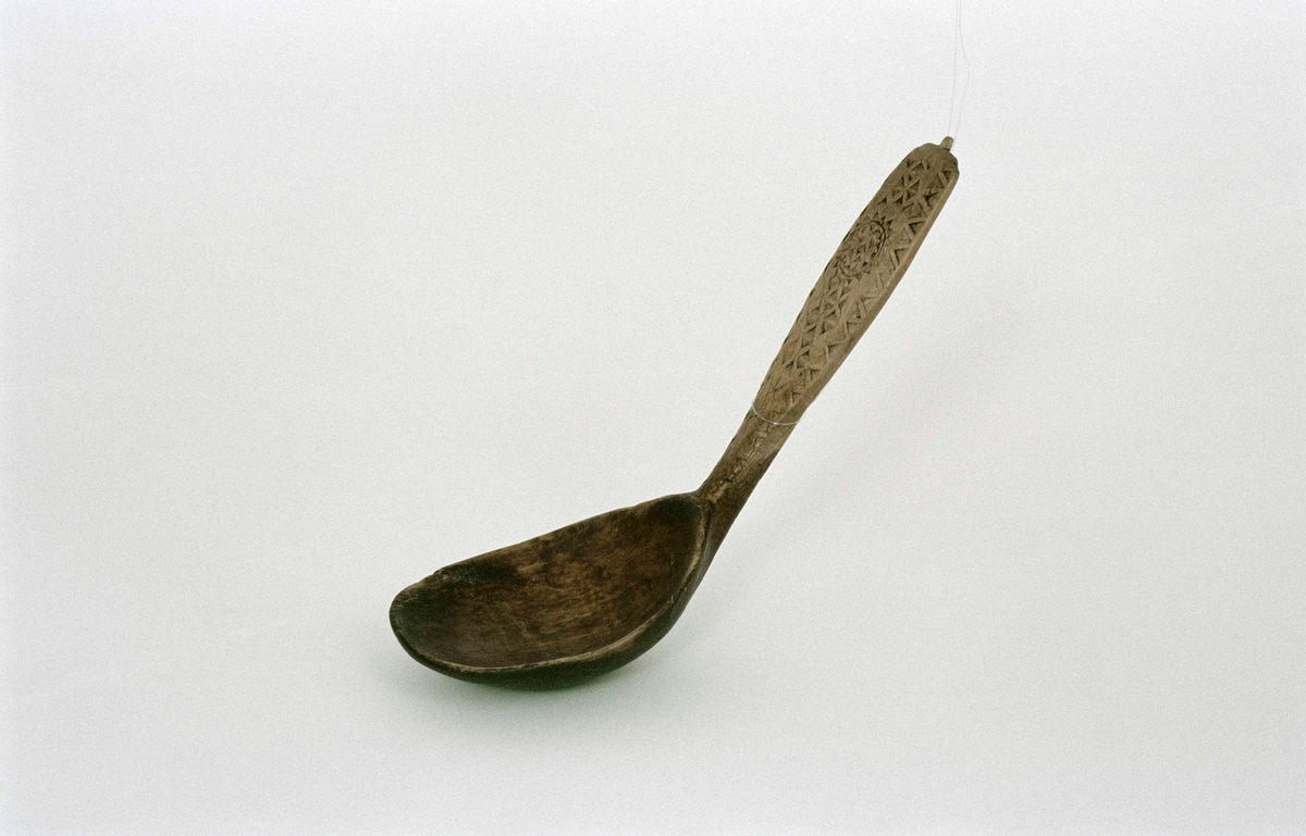 Sked av lövträ med riklig karvsnittsornamentik på skaftet. Djupt blad, böjt mot skaftet. Spår av grön bemålning på bladet.