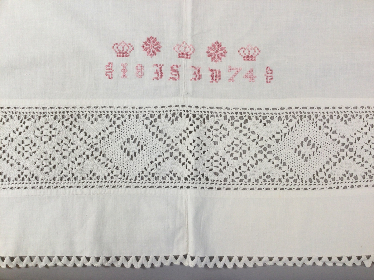 Överlakan i tuskaftad bomullsväv, till brud. Två våder, hopsytt för hand, maskinfållat i över- och nederkant. Infälld knypplad bård, vitt bomullsgarn, ca 11 cm bred, drygt 7 cm från överkanten, romber och hjärtan. Kantat med gansband upptill och 30 cm ned på var sida. Monogram: 18 IS ID 74, kronor och stjärnor, broderi; korssöm, årtalet broderat i fyrkanter påminnande om myrgång, rosa bomullsgarn.