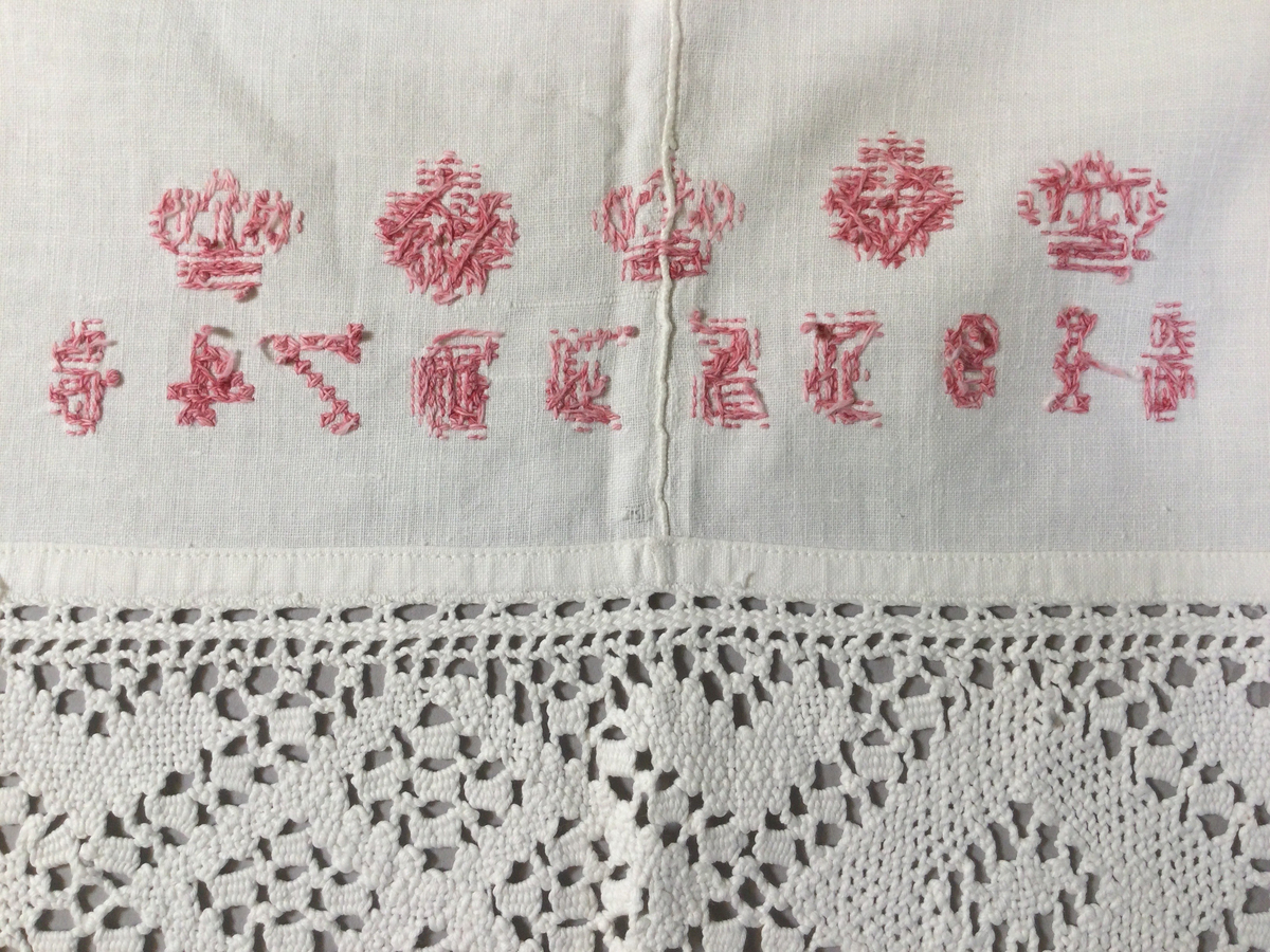 Överlakan i tuskaftad bomullsväv, till brud. Två våder, hopsytt för hand, maskinfållat i över- och nederkant. Infälld knypplad bård, vitt bomullsgarn, ca 11 cm bred, drygt 7 cm från överkanten, romber och hjärtan. Kantat med gansband upptill och 30 cm ned på var sida. Monogram: 18 IS ID 74, kronor och stjärnor, broderi; korssöm, årtalet broderat i fyrkanter påminnande om myrgång, rosa bomullsgarn.