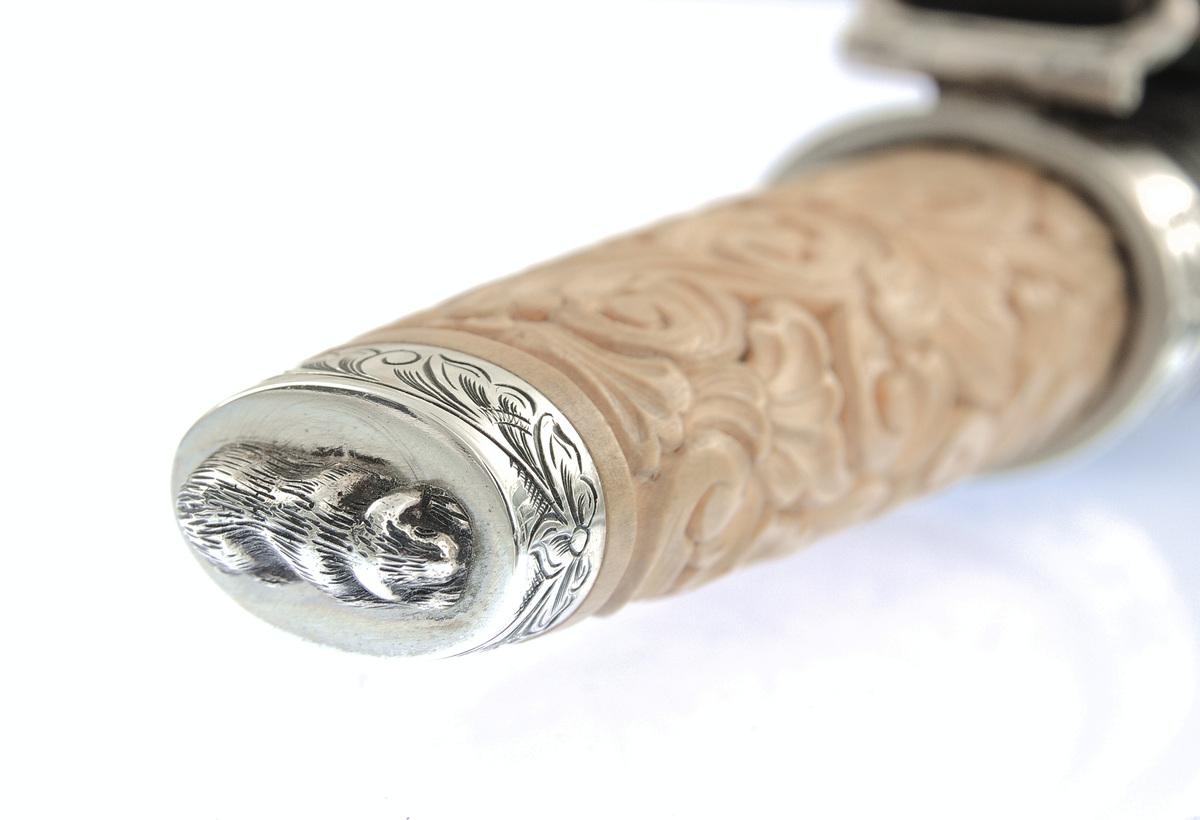 Kniv med utskore skaft og gravera holkar, på toppen ein støypt hund.
Slire av horn, utskoren på begge sider, holkar av sylv som er gravera.