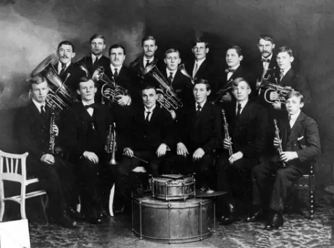 Hauge Musikkorps fra Rolvsøy med sin dirigent Oskar Lier. Bildet er tatt i et fotoatelier. Navneliste% (opplysninger fra Leiv B. Kristiansen 1992).