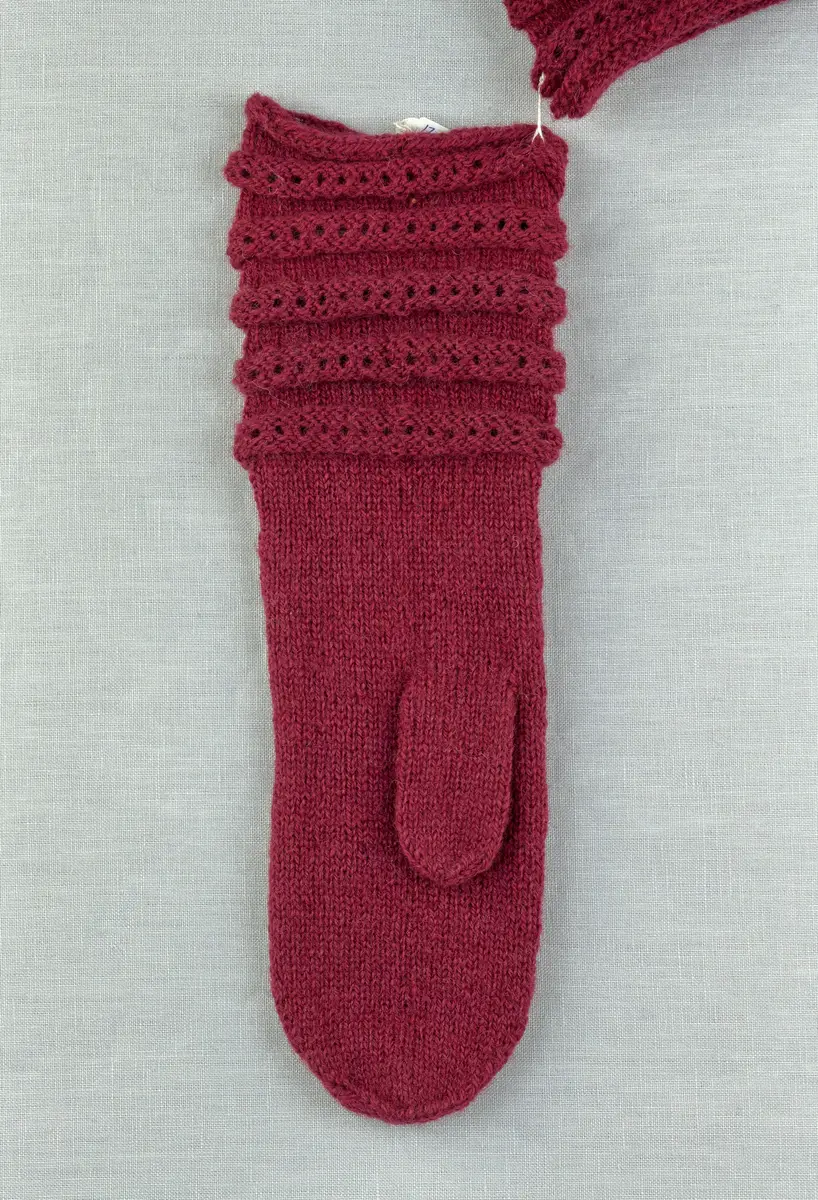 Ett par stickade tumvantar, med ränder i avigstickning och ett hålvarv mitt på varje rand på mudd/krage och vante i påfågelmönstrad spets-stickning i 2-trådigt ullgarn. Vanten har instickad tumme och toppen är formad med bandhoptagning.