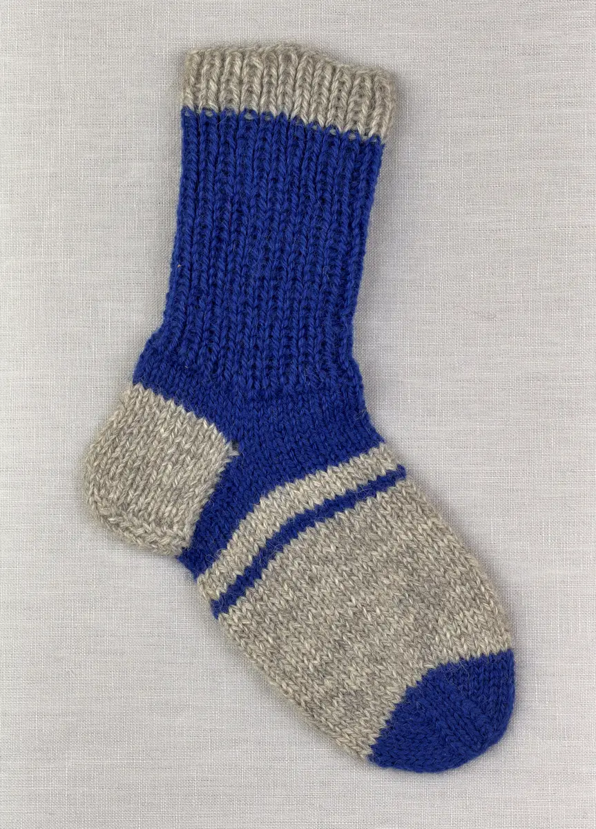 En stickad socka, med randig mudd i 1-1 resår (1 rät och 1 avig maska) och randig fot i slätstickning, av 3-trådigt Sportgarn i ull.
Gjord efter Bergå beskrivning D 315 från Bergå Färgeri & Ullspinneri, Stora Skedvi, Dalarna.