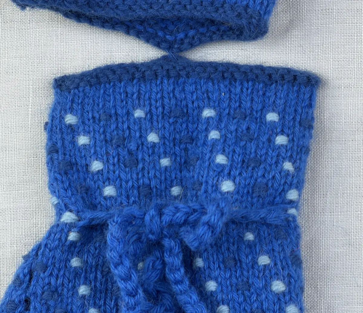 Ett par stickade damvantar, i slätstickning med nockor på vantens insida, av 3-trådigt sportgarn i ull. Nockornas/knutarnas "baksida" bildar ett prickmönster på vantens utsida. 
Vanten har två garntofsar med virkad snodd, kiltumme och är avslutad med bandhoptagning i toppen.