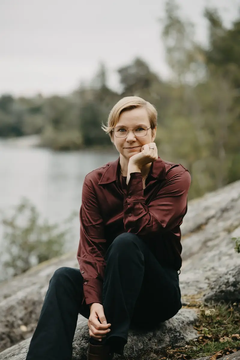 Kvinna i rödbrun tröja och svarta byxor sitter på en klipphäll med vatten och skog i bakgrunden.