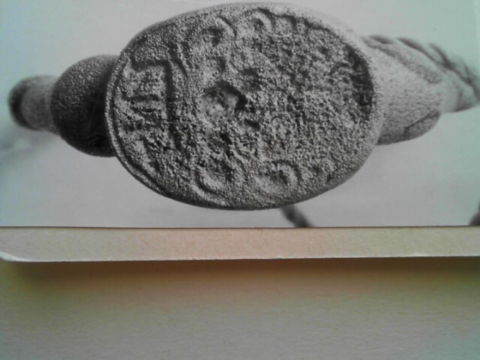 Messing. Tinn ?. Oval ring. Oval signetplate med utydelig mønster. Spor av rød lakk (?) i platen. Ryggen av platen har riller. Muligens overtrekk av tinn (?) på ringen.