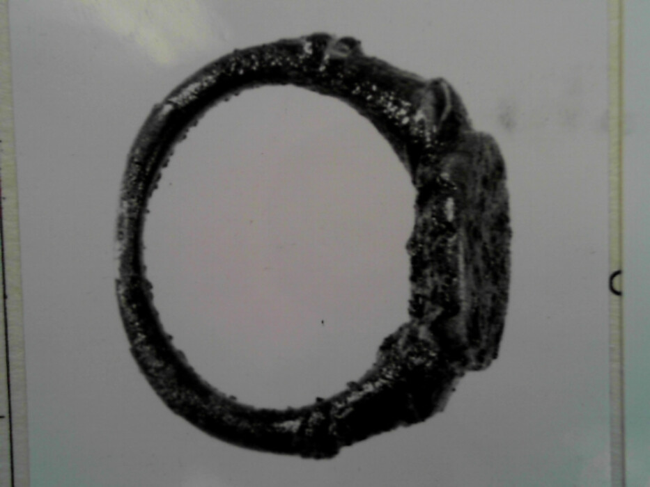 Messing. Tinn ?. Oval ring. Oval signetplate med utydelig mønster. Spor av rød lakk (?) i platen. Ryggen av platen har riller. Muligens overtrekk av tinn (?) på ringen.