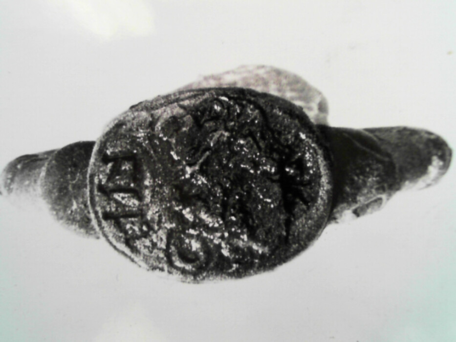 Messing. Tinn ?. Oval ring. Oval signetplate med utydelig mønster. Spor av rød lakk (?) i platen. Ryggen av platen har riller. Muligens overtrekk av tinn (?) på ringen.