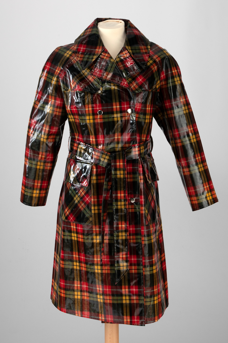 Regnfrakk med belte og hatt. Skotskrutet i fargene svart, grønt, rødt, gult og hvitt. Frakken er lettskrådd med midtsøm i ryggen og splitt nederst, innskutte sidestykker i hele høyden og knapping foran med trykknapper i hvitt metall. Løst bærestykke i ryggen og på hver side foran. Nedbrettet, ganske bred krage med spisser. Isydde, rette ermer. På hvert forstykke en påstukket stor lomme med klaff. Knyttebelte gjennom stropper i sidene. Regnhatt med høy pull, satt sammen av seks stoffstykker. Dobbeltsydd brem med 10cm bredde.