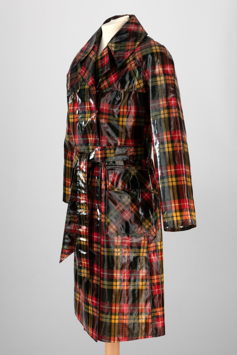 Regnfrakk med belte og hatt. Skotskrutet i fargene svart, grønt, rødt, gult og hvitt. Frakken er lettskrådd med midtsøm i ryggen og splitt nederst, innskutte sidestykker i hele høyden og knapping foran med trykknapper i hvitt metall. Løst bærestykke i ryggen og på hver side foran. Nedbrettet, ganske bred krage med spisser. Isydde, rette ermer. På hvert forstykke en påstukket stor lomme med klaff. Knyttebelte gjennom stropper i sidene. Regnhatt med høy pull, satt sammen av seks stoffstykker. Dobbeltsydd brem med 10cm bredde.