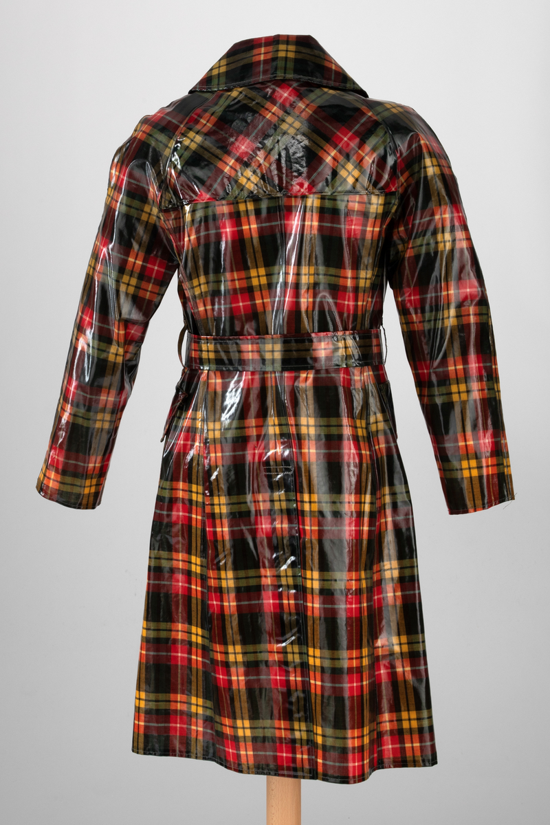 Regnfrakk med belte og hatt. Skotskrutet i fargene svart, grønt, rødt, gult og hvitt. Frakken er lettskrådd med midtsøm i ryggen og splitt nederst, innskutte sidestykker i hele høyden og knapping foran med trykknapper i hvitt metall. Løst bærestykke i ryggen og på hver side foran. Nedbrettet, ganske bred krage med spisser. Isydde, rette ermer. På hvert forstykke en påstukket stor lomme med klaff. Knyttebelte gjennom stropper i sidene. Regnhatt med høy pull, satt sammen av seks stoffstykker. Dobbeltsydd brem med 10cm bredde.