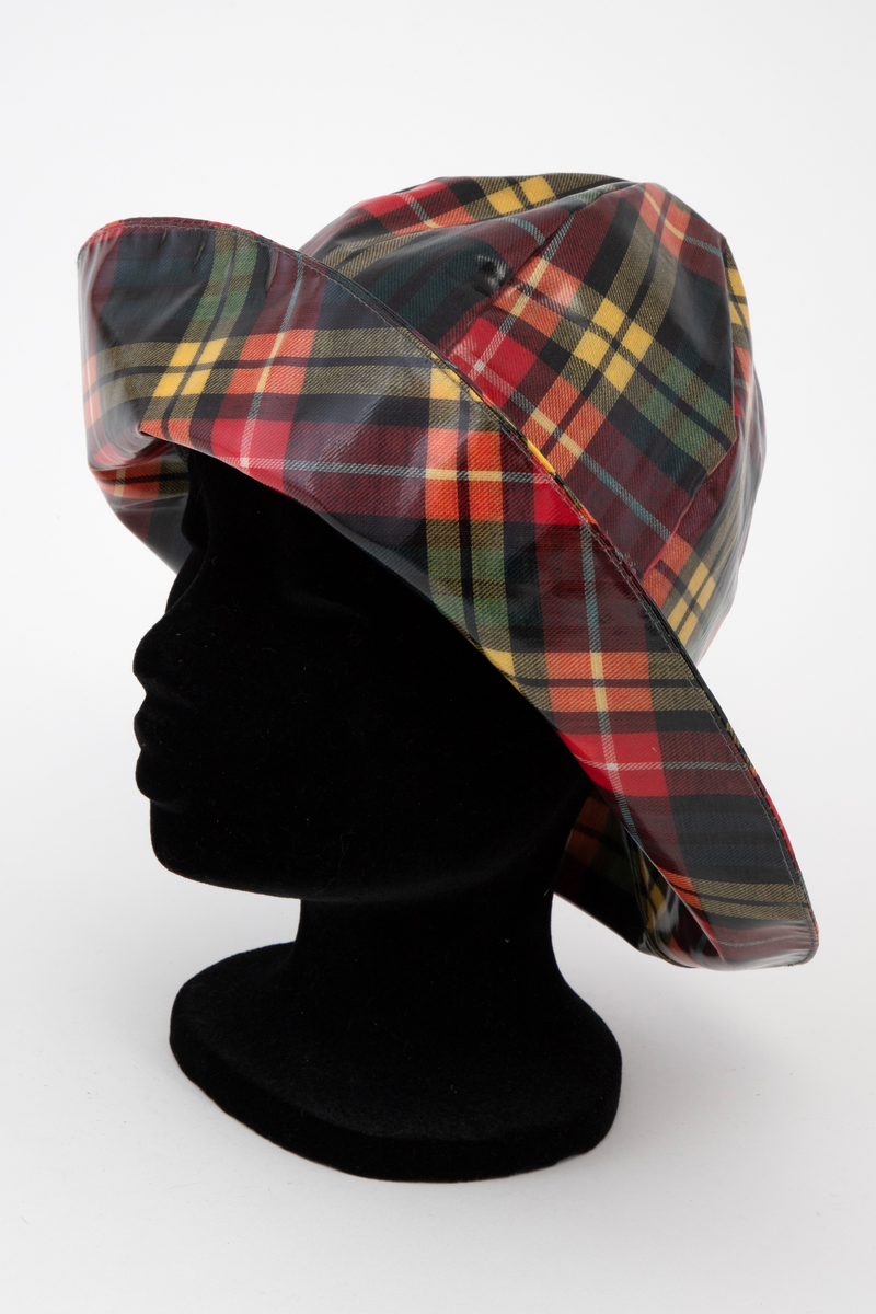 Regnfrakk med belte og hatt. Skotskrutet i fargene svart, grønt, rødt, gult og hvitt. Frakken er lettskrådd med midtsøm i ryggen og splitt nederst, innskutte sidestykker i hele høyden og knapping foran med trykknapper i hvitt metall. Løst bærestykke i ryggen og på hver side foran. Nedbrettet, ganske bred krage med spisser. Isydde, rette ermer. På hvert forstykke en påstukket stor lomme med klaff. Knyttebelte gjennom stropper i sidene. Regnhatt med høy pull, satt sammen av seks stoffstykker. Dobbeltsydd brem med 10cm bredde.