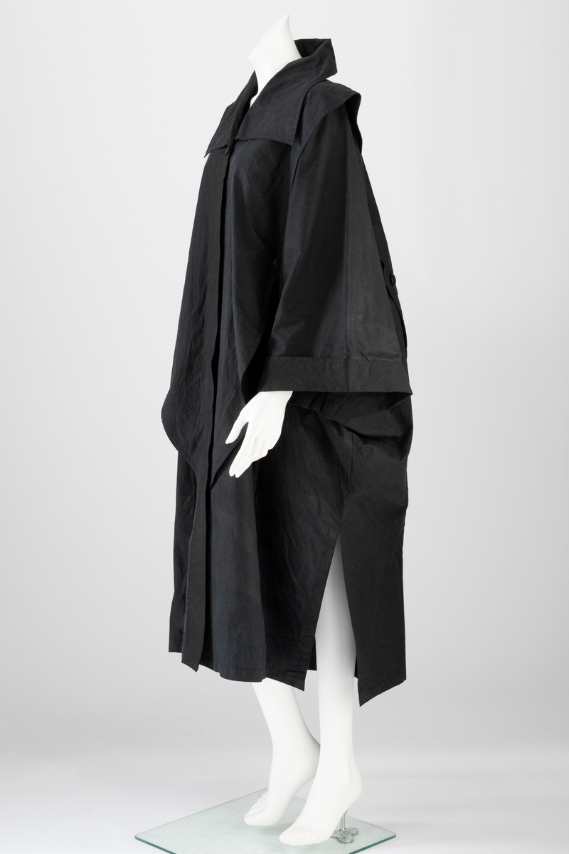 Svart regnkappe i kimonofasong med oppbrett på ermene og stor, rett, utbrettet krage. Kappen er fôret med svart, tynt bomullsstoff. Løst stoffstykke foran som går ned i en spiss, sydd til stolpen og festet med en knapp i ytterkant. Bak er det løse stykket sydd ned til en søm 5,5cm fra midt bak på begge sider av midten. Kappen lukkes med trykknapper og en knapp og knapphull ved halsen. Rygg og forstykke er sydd i ett stykke. Krage, oppbrett på ermene og stoffstykket foran er ensfarget svart. Stykket bak har bølgeformet dekor (stofftrykk). Tilhørende ensfarget svart, sirkelrund, flat lue. Oppå pullen er det sydd fast to løse, doble kanter, festet med en knapp i yttersiden.