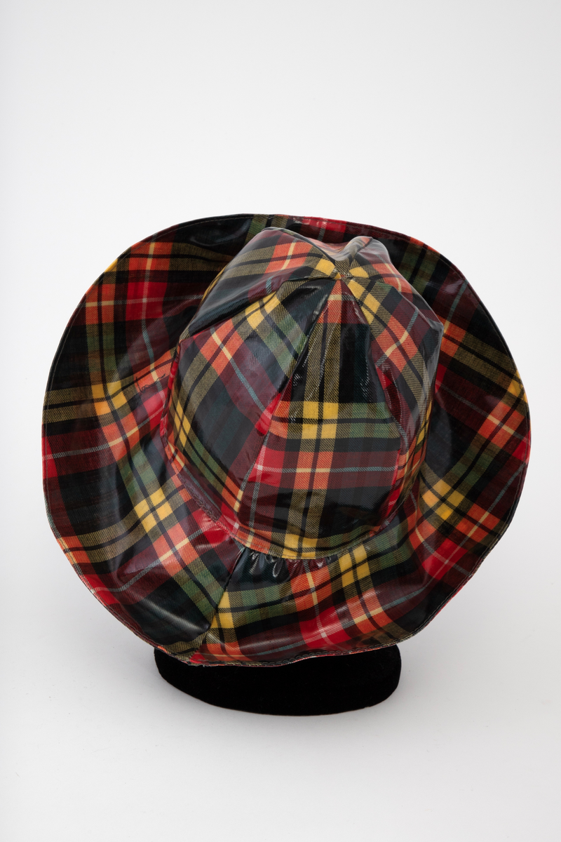 Regnfrakk med belte og hatt. Skotskrutet i fargene svart, grønt, rødt, gult og hvitt. Frakken er lettskrådd med midtsøm i ryggen og splitt nederst, innskutte sidestykker i hele høyden og knapping foran med trykknapper i hvitt metall. Løst bærestykke i ryggen og på hver side foran. Nedbrettet, ganske bred krage med spisser. Isydde, rette ermer. På hvert forstykke en påstukket stor lomme med klaff. Knyttebelte gjennom stropper i sidene. Regnhatt med høy pull, satt sammen av seks stoffstykker. Dobbeltsydd brem med 10cm bredde.